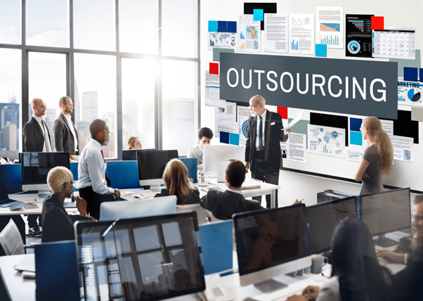 A small team collaborating around laptops in a modern open-plan office, with a wall display showing the word OUTSOURCING — the central theme of small-business outsourcing.